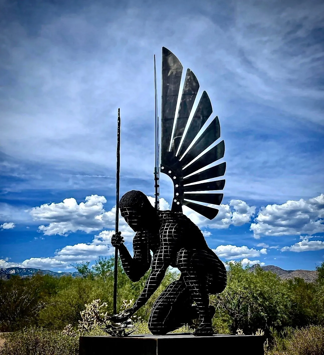 A dark angel of steel slices kneeling with a spear, a fanned wing rising behind, under a blue sky